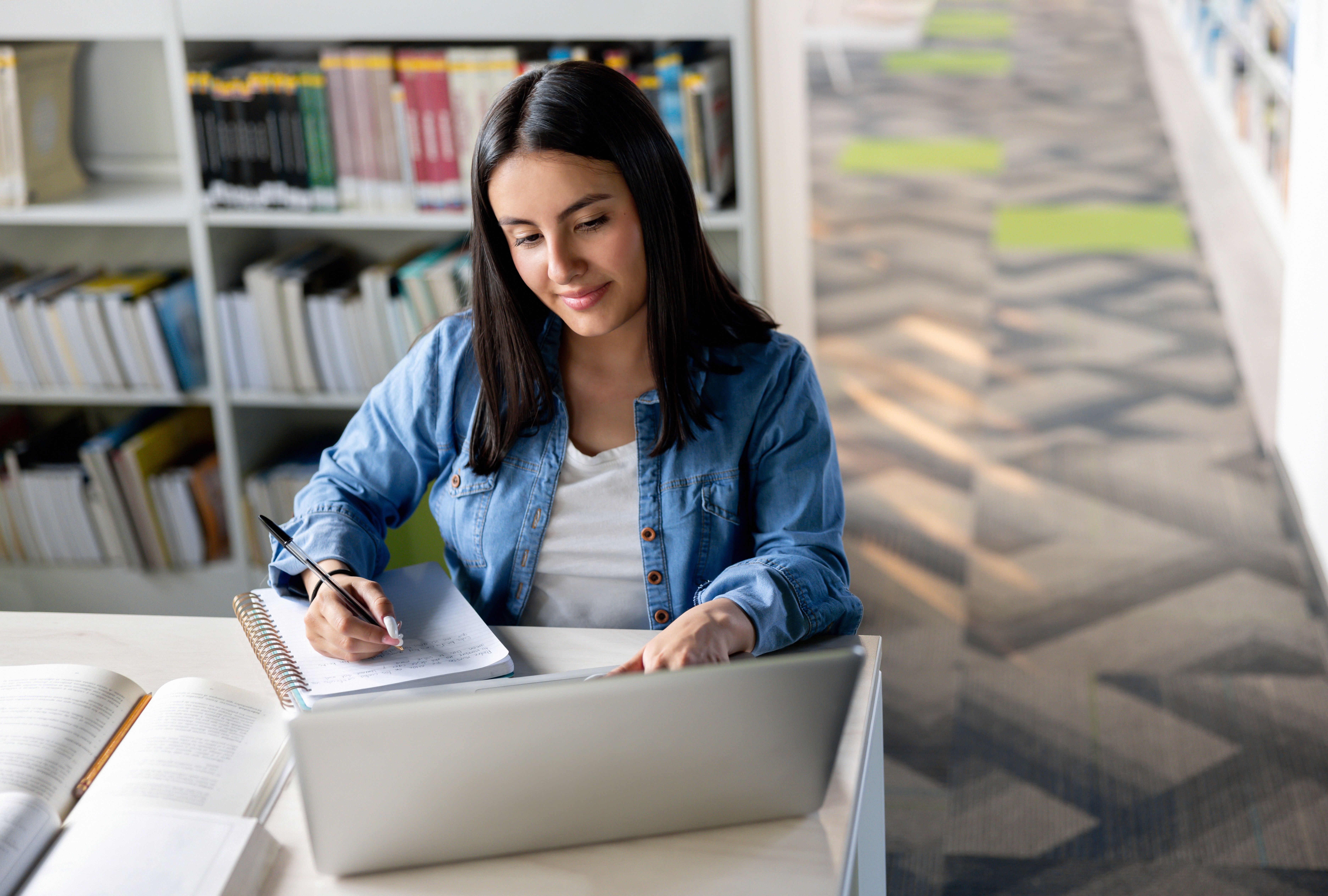 A Female Student Studying At Library Using Laptop And Writing In Notebook