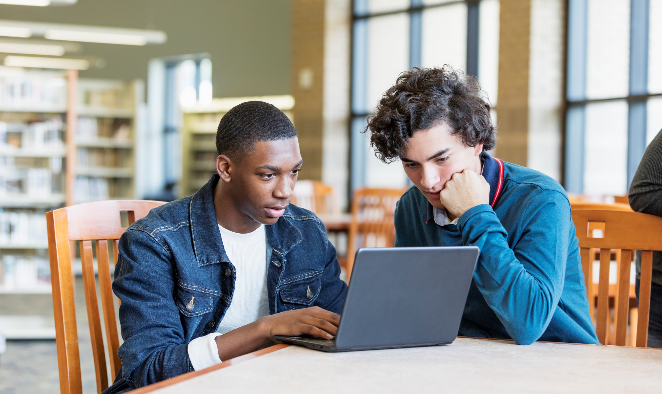 Two Male Teens Looking Over Laptop Together in Library