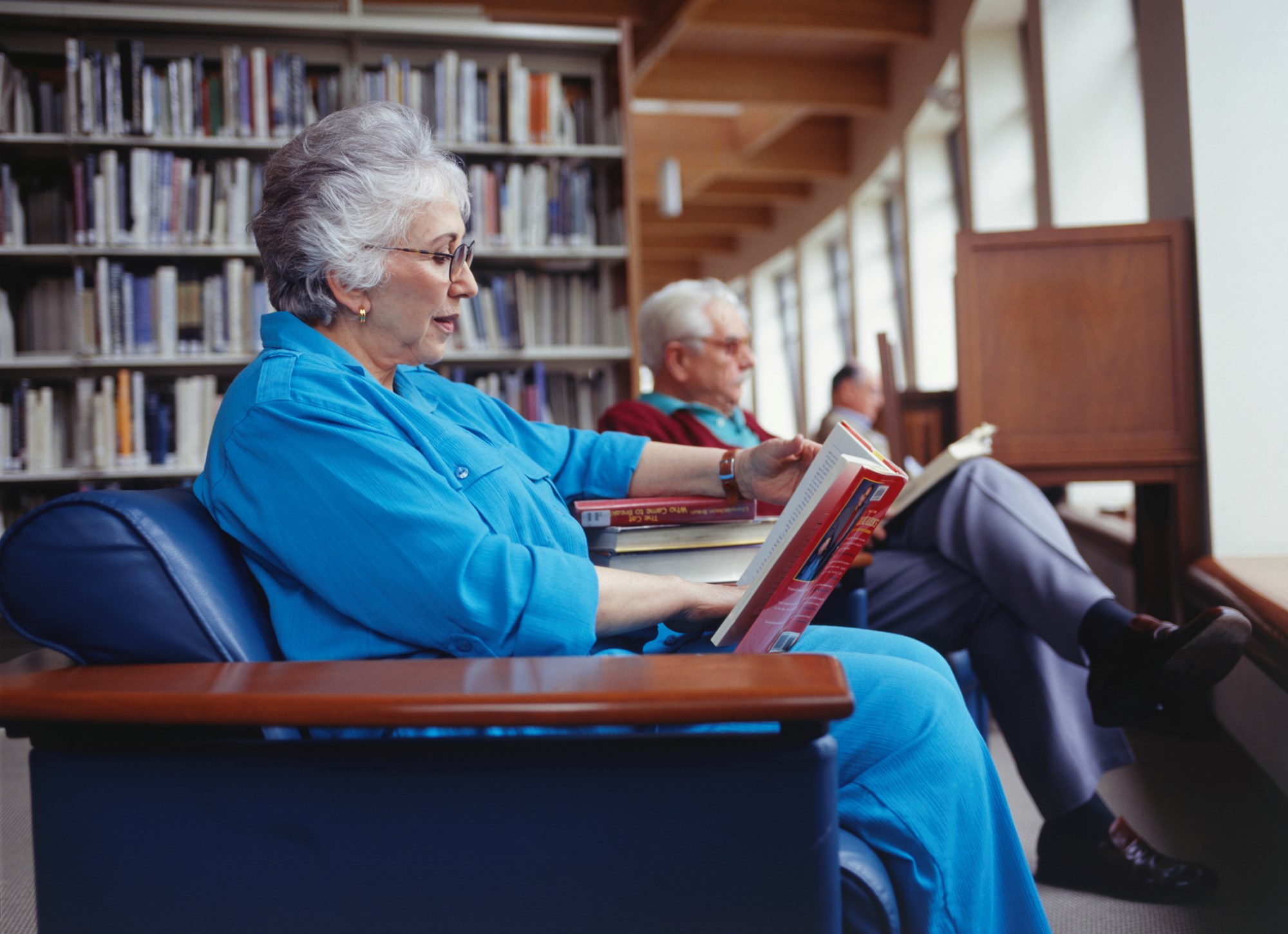 A photo of two older adults sitting in chairs and reading in a library.