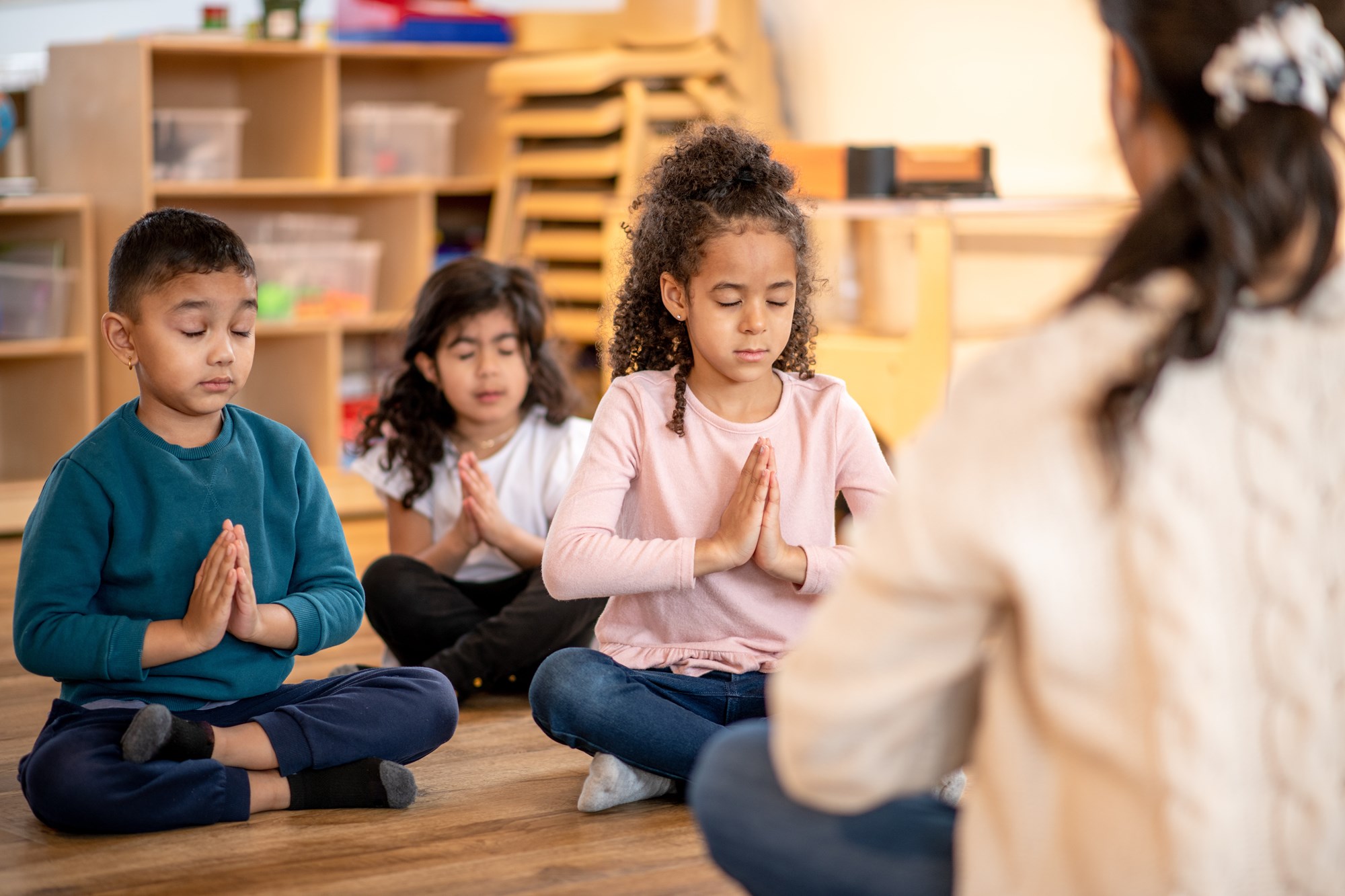 A photo of children sitting on the floor of a classroom, facing an adult also sitting on the floor.