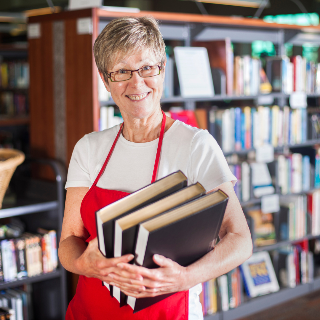 An older woman in a red apron stands in a library and holds three books.