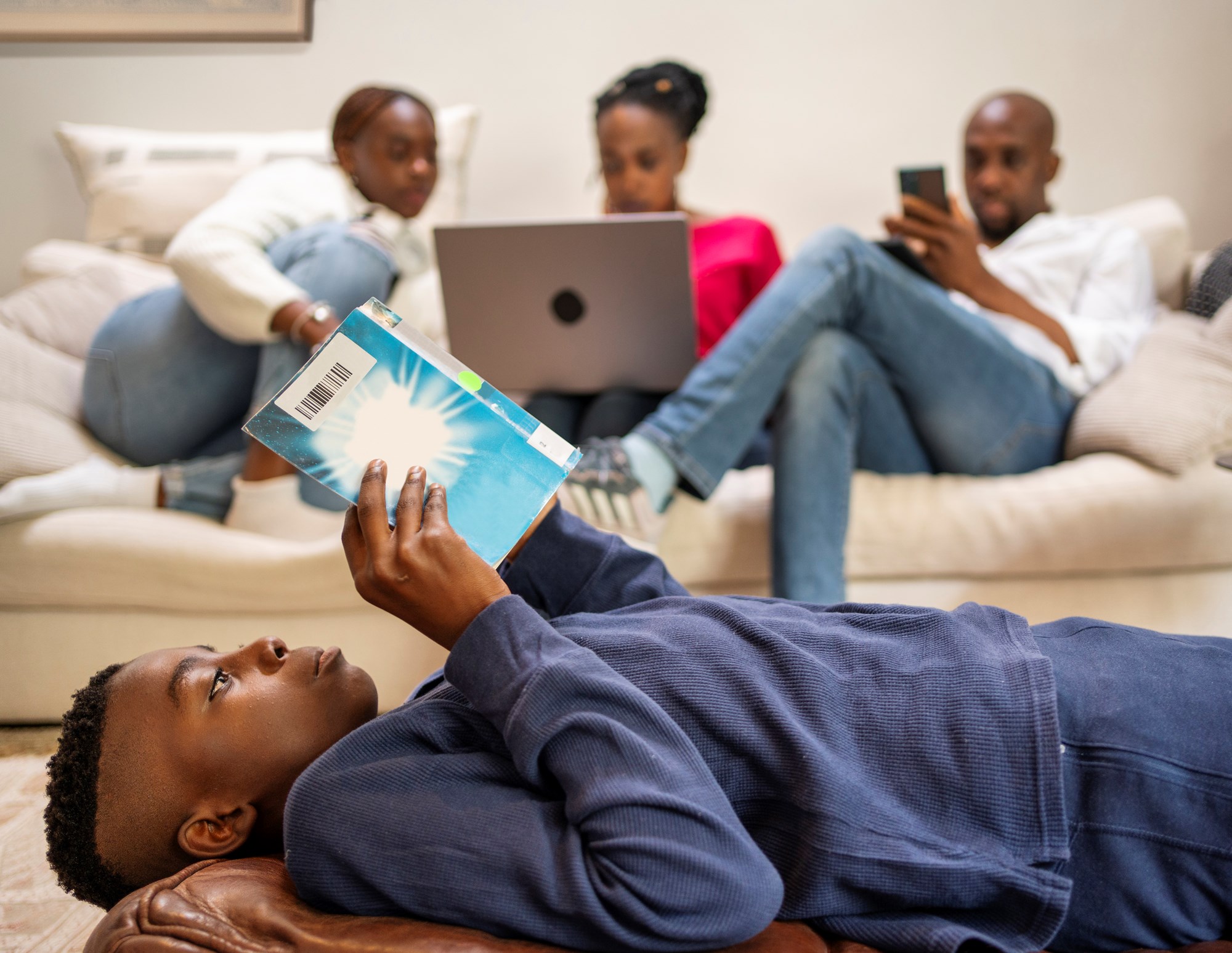 Boy on Floor Reading Book with Family Relaxing on Sofa at Home