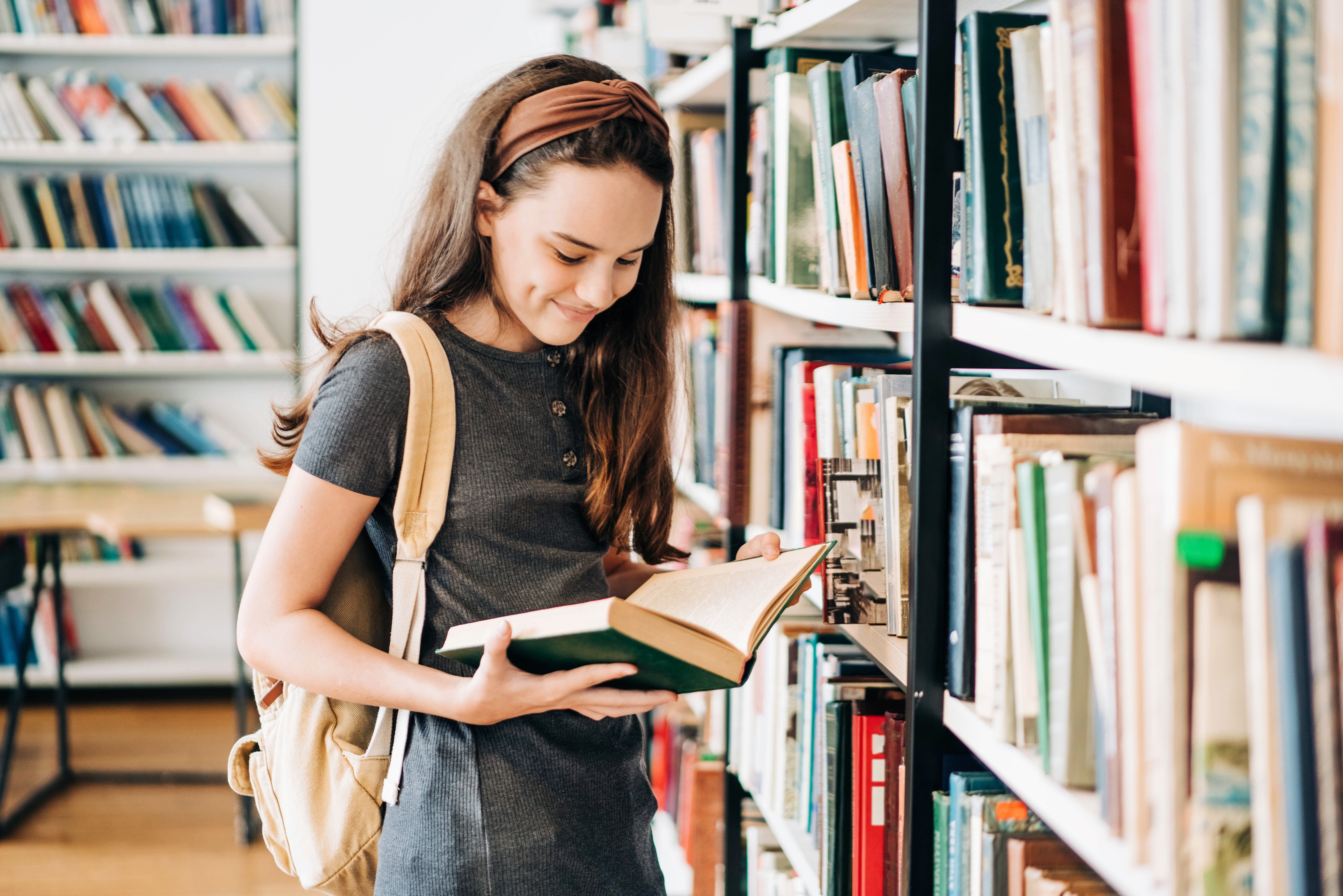 A Smiling Teenage Girl with a Backpack Selects a Book From Library