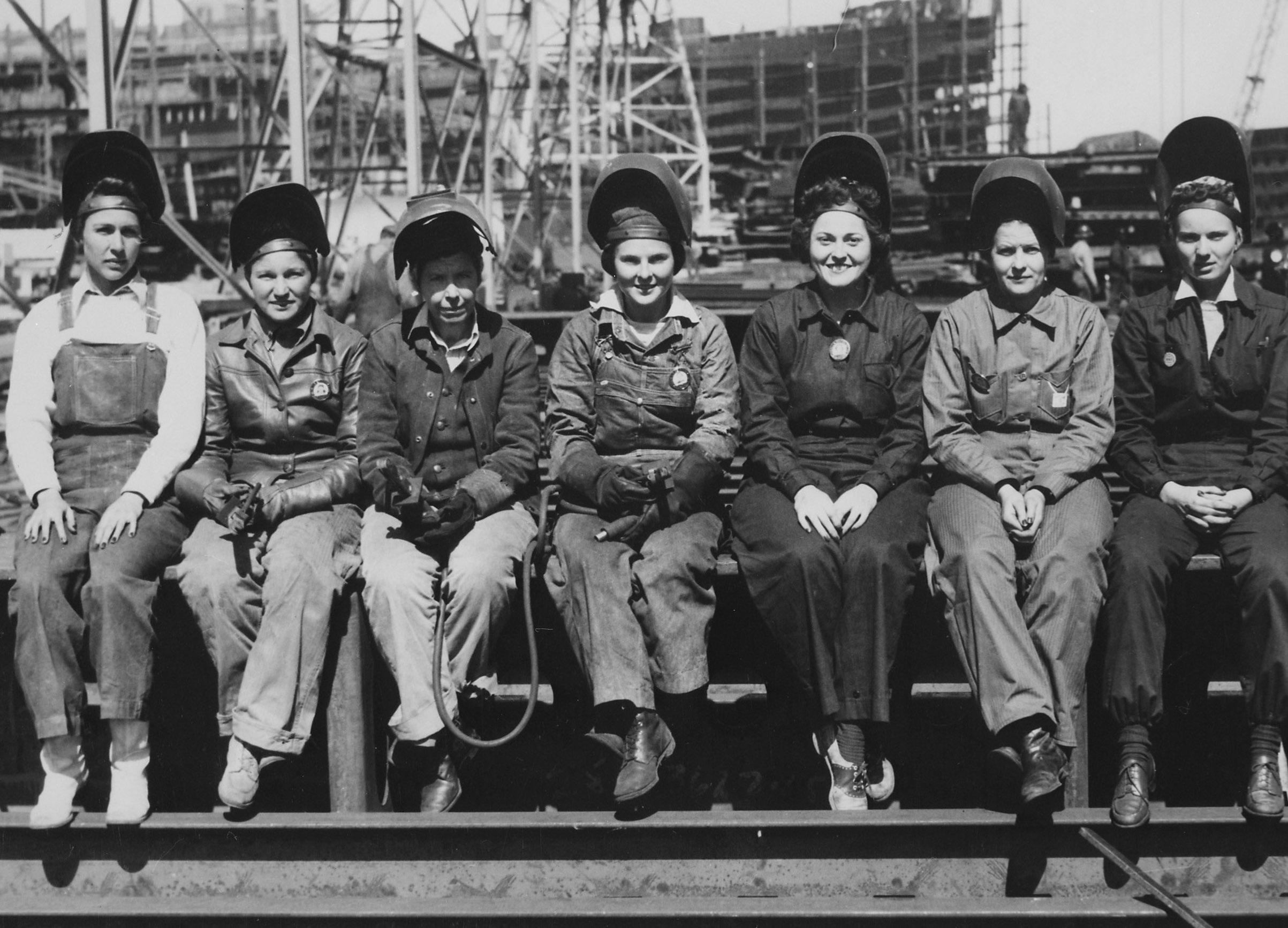 Seven women in welding gear sit side by side at a WWII shipyard