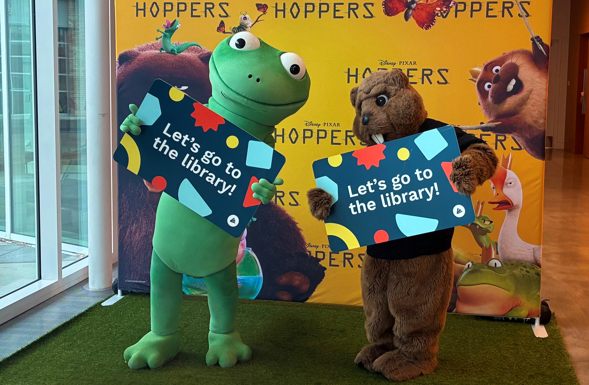 People in lizard and beaver costumes holding up giant library cards in front of Hoppers movie banner.