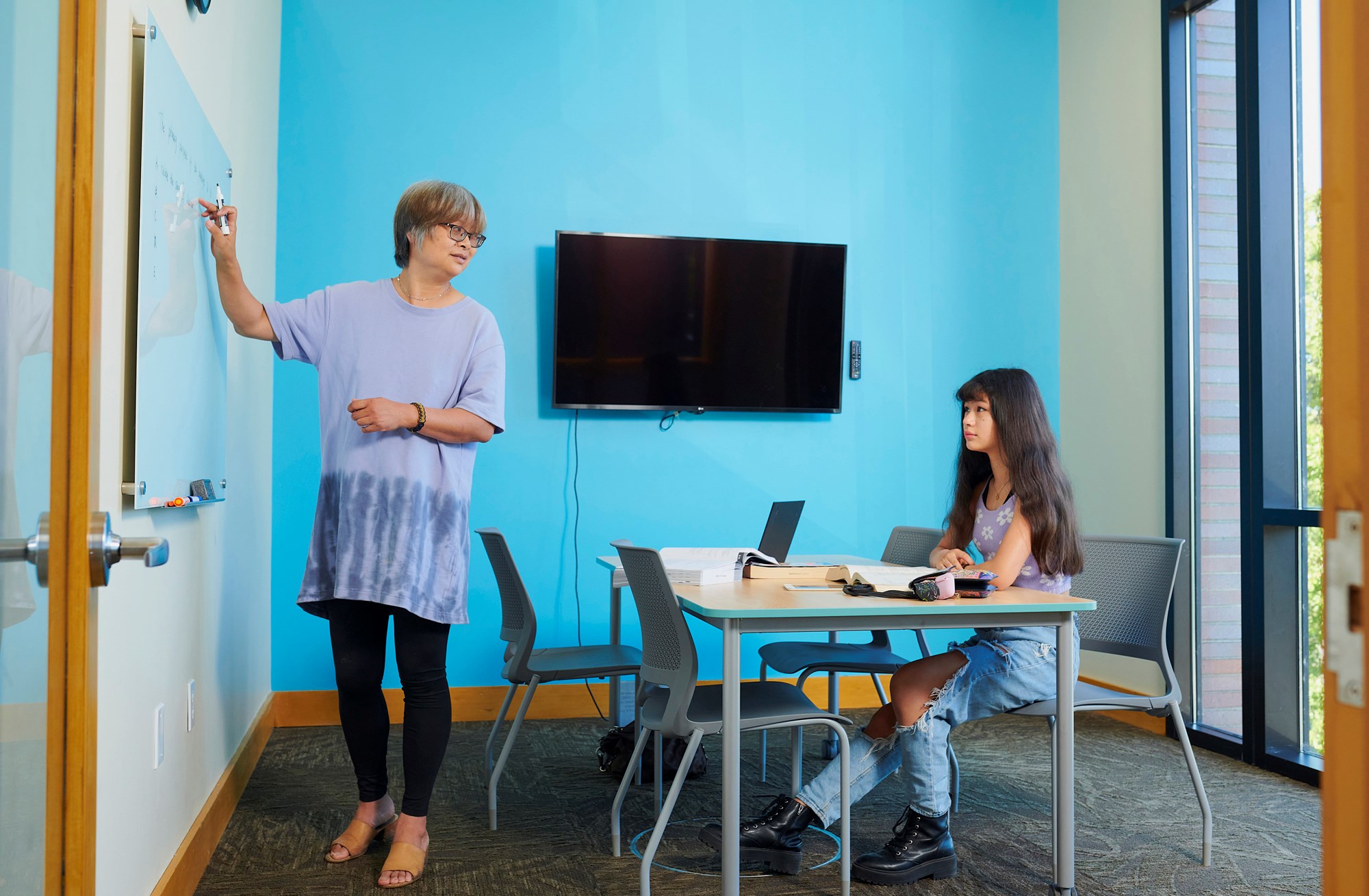 Two people using a Study Cube in a library, a small room with a table, TV monitor, and whiteboard.