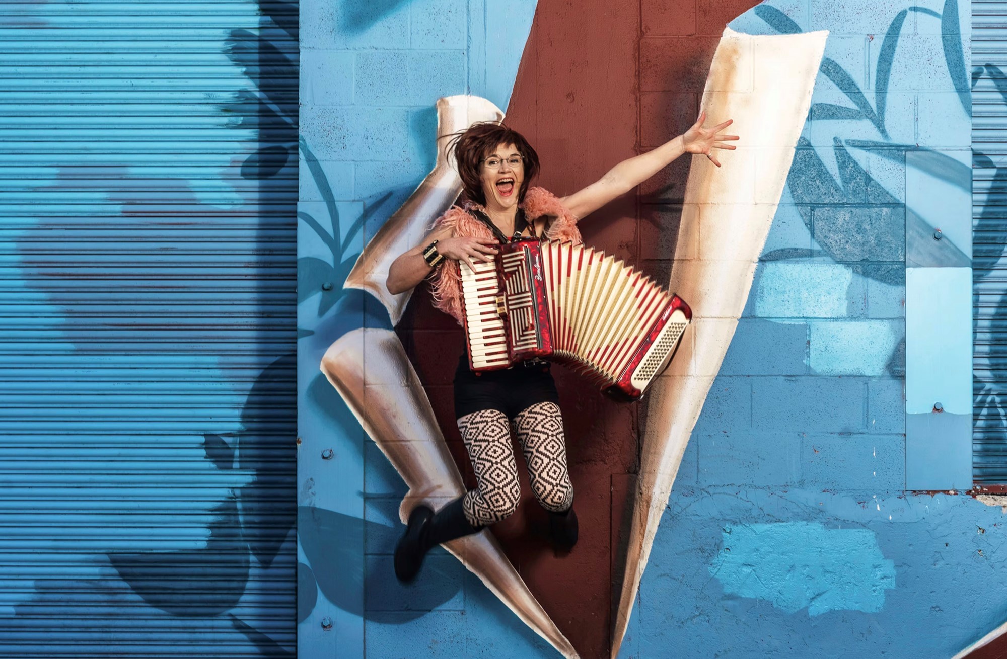 Woman with accordion jumping in front of blue and red mural.