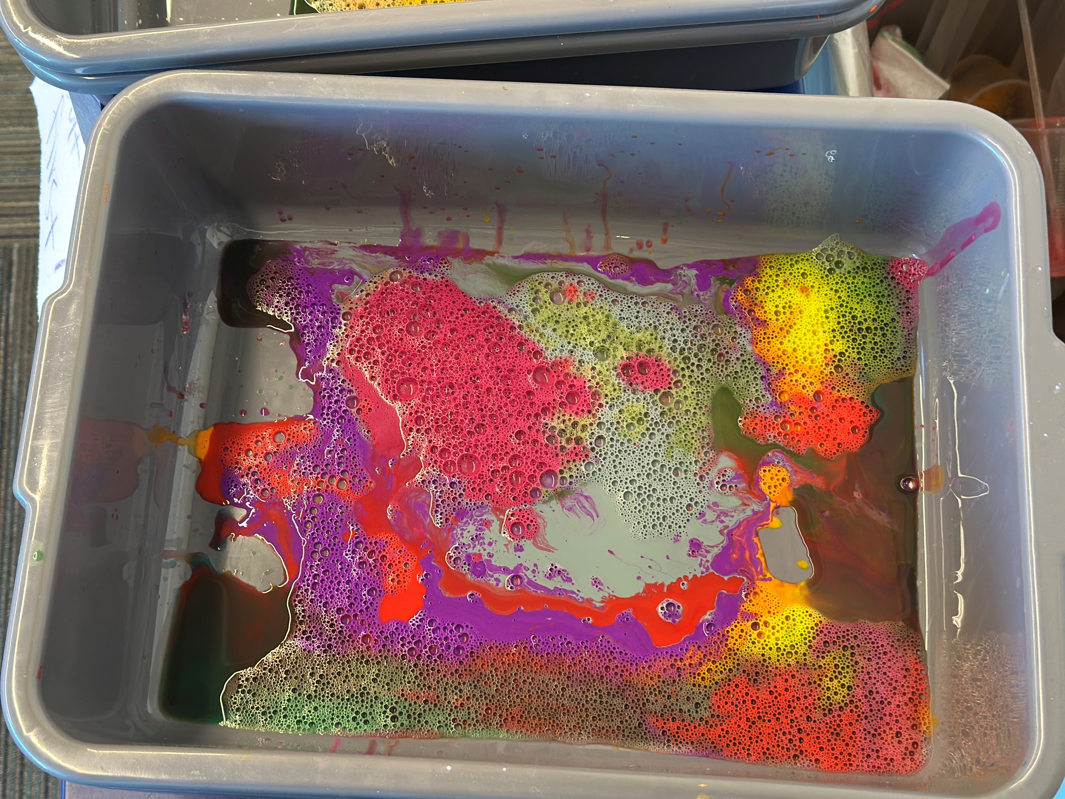 A gray bussing bin with filmy water and swirls of fuchsia, yellow, turquoise, and blue.