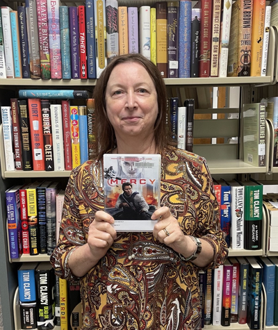 Library worker standing in front of bookshelf holding Mercy DVD.
