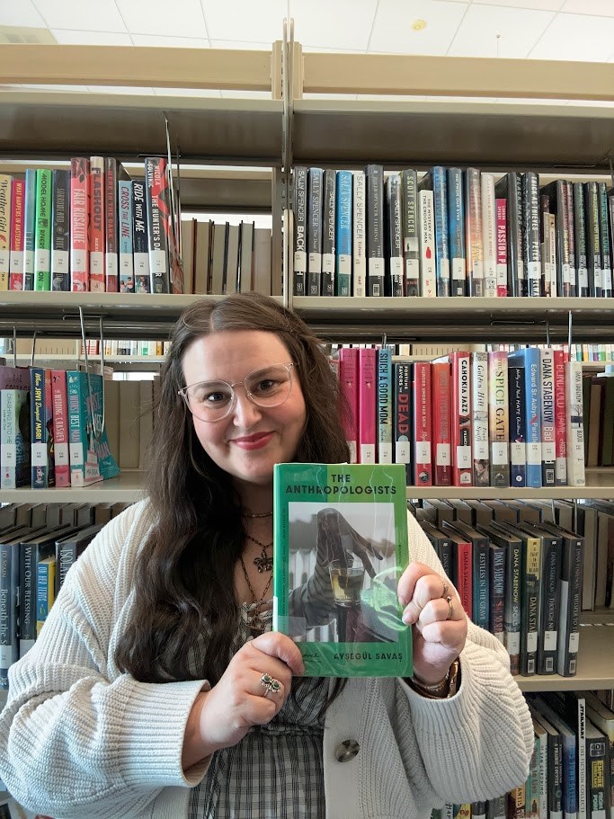 Rachael standing in front of a bookshelf holding a copy of The Anthropologists by Ayşegül Savaş