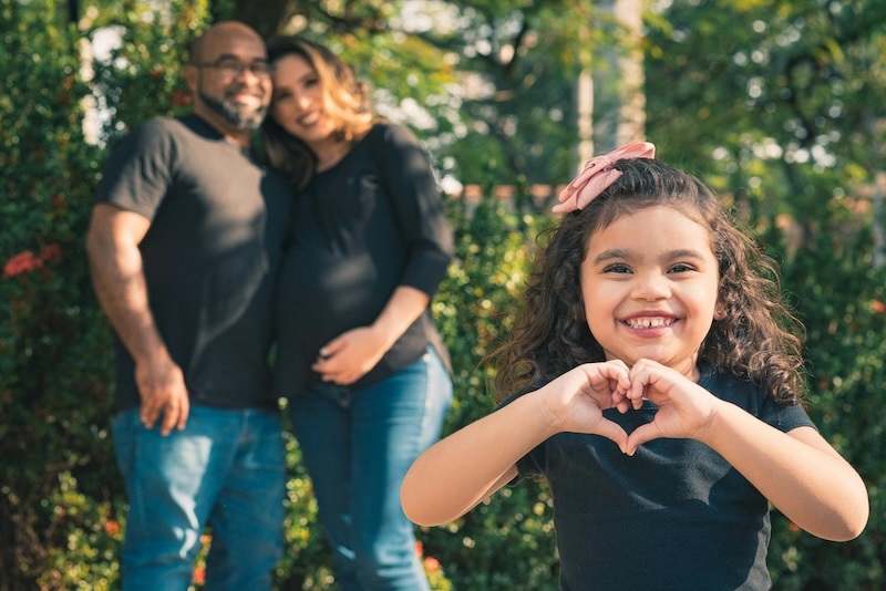 A young girl making a heart shape with her hands at the viewer. Her parents are smiling in the background.