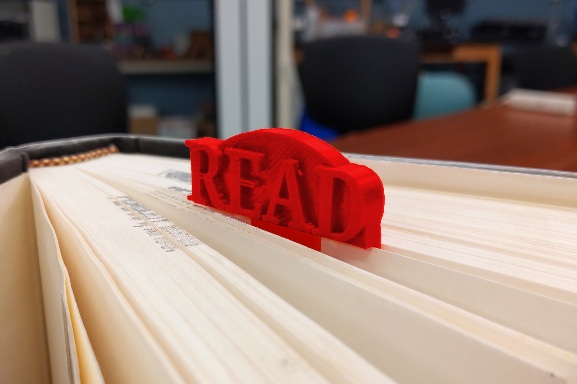 3D-printed bookmark with the word "READ" embossed on it, clipped onto a page of a book