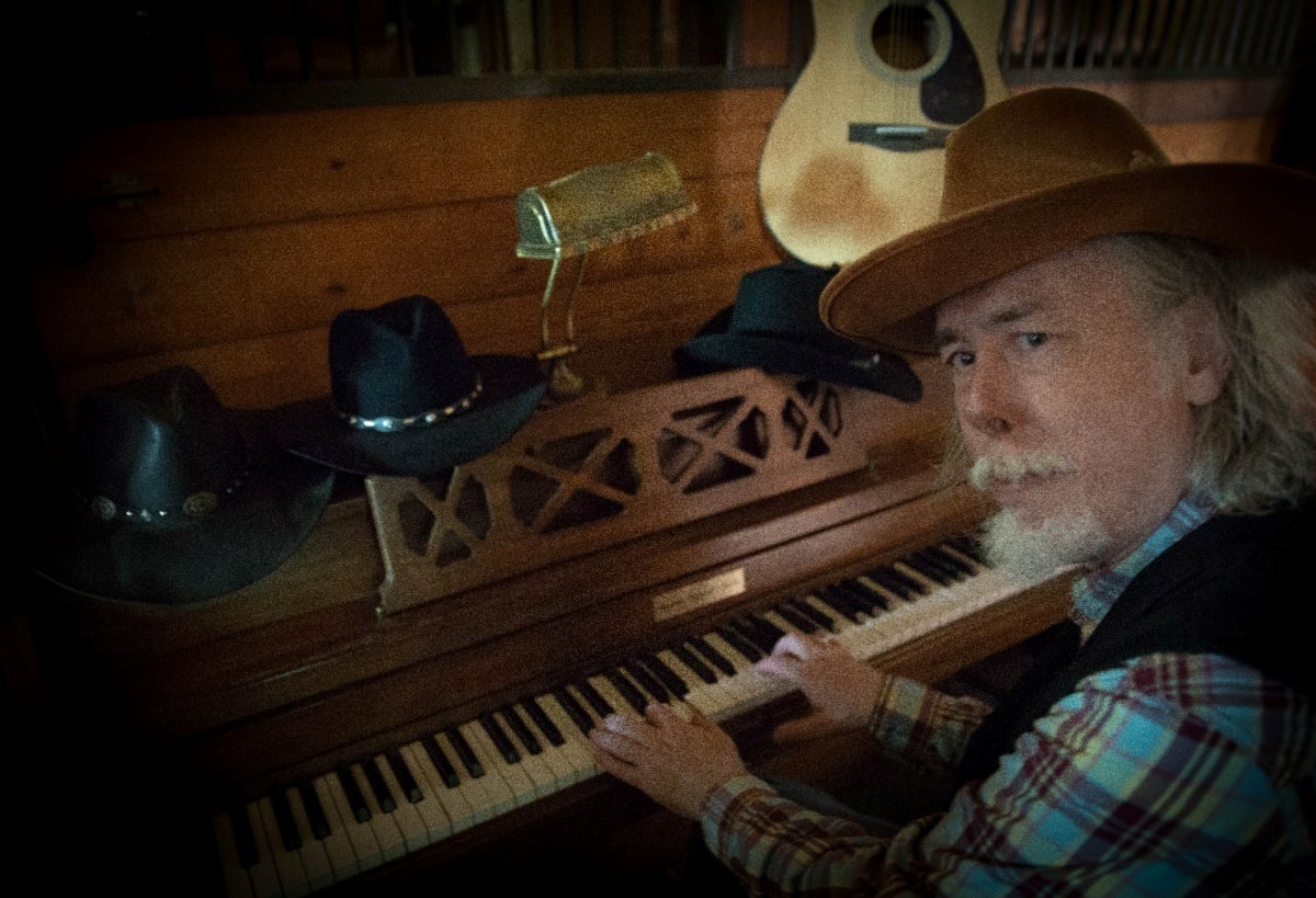Picture of a man with grey hair and a grey beard sitting at an upright piano.