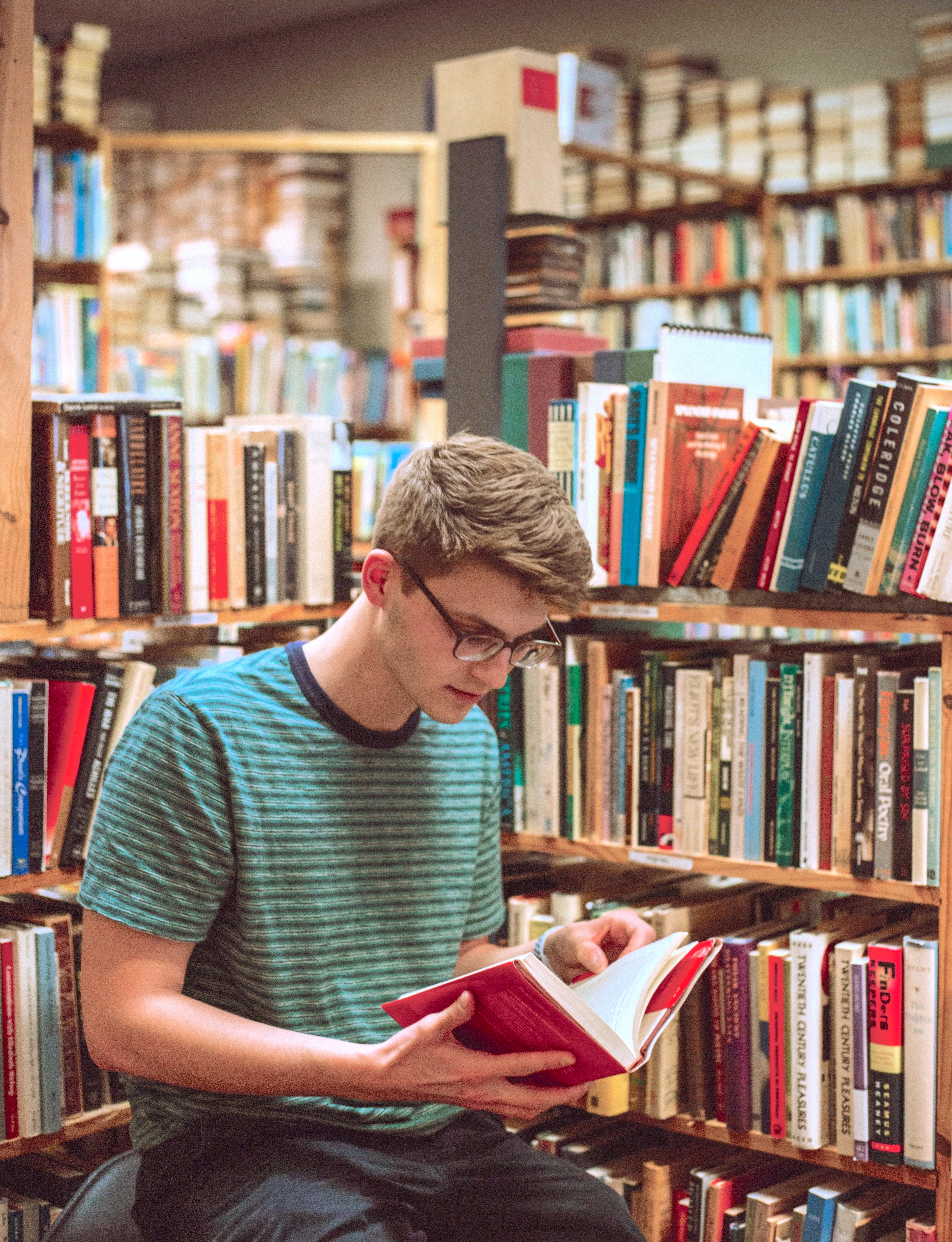 Picture of a teen boy sitting by a bookshelf reading a book.
