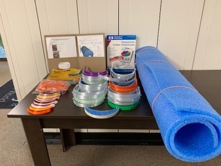 A picture of a brown table with 3D printer face maks components, a blue roll of foam padding,and clear plastic face shields.