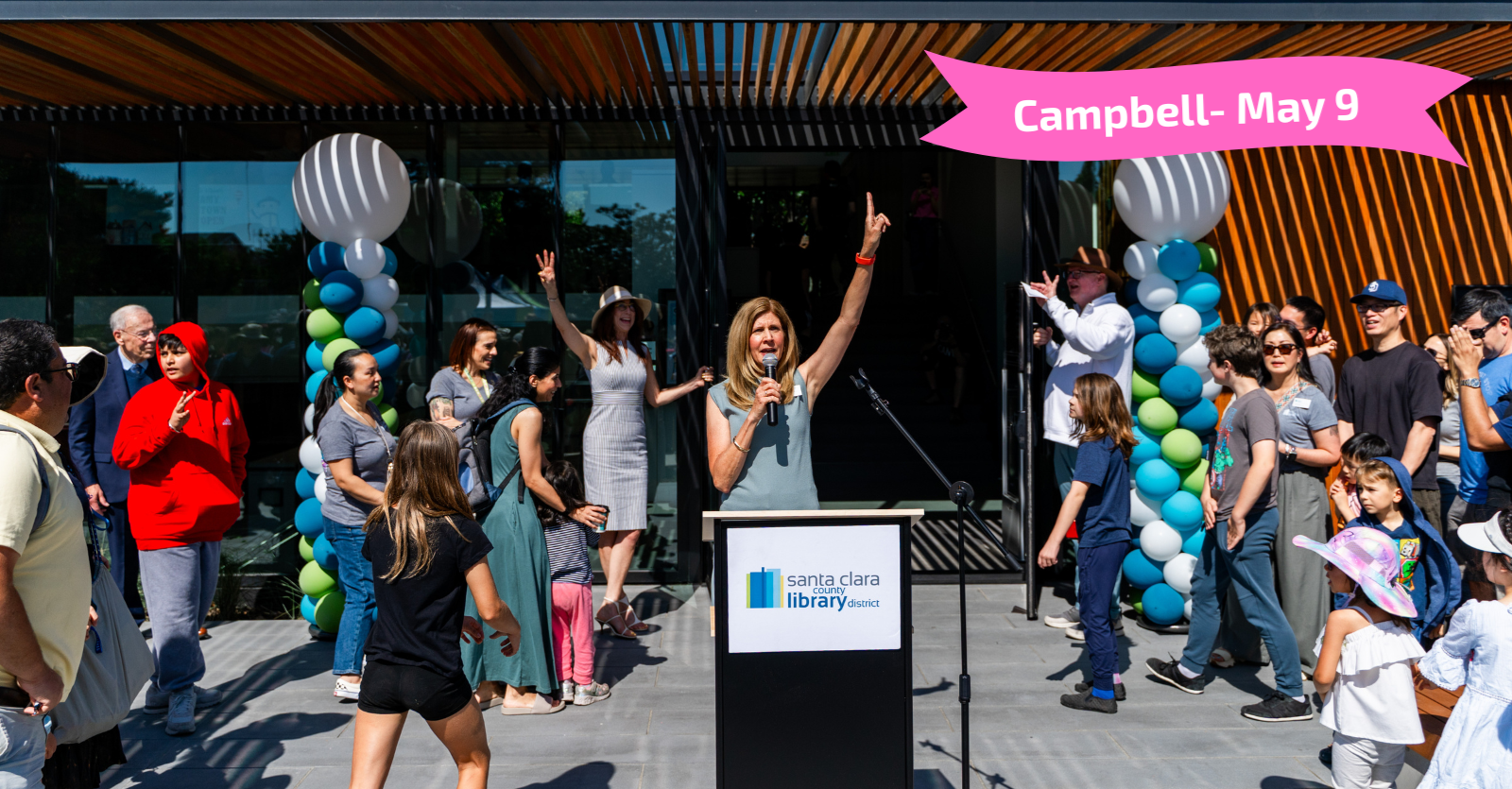 Image from opening of library. County Librarian Jennifer Weeks raises her left arm to announce that the library is open as a crowd of onlookers begins to make their way toward the entrance. There are two pilons of colorful balloons and event date, May 9.