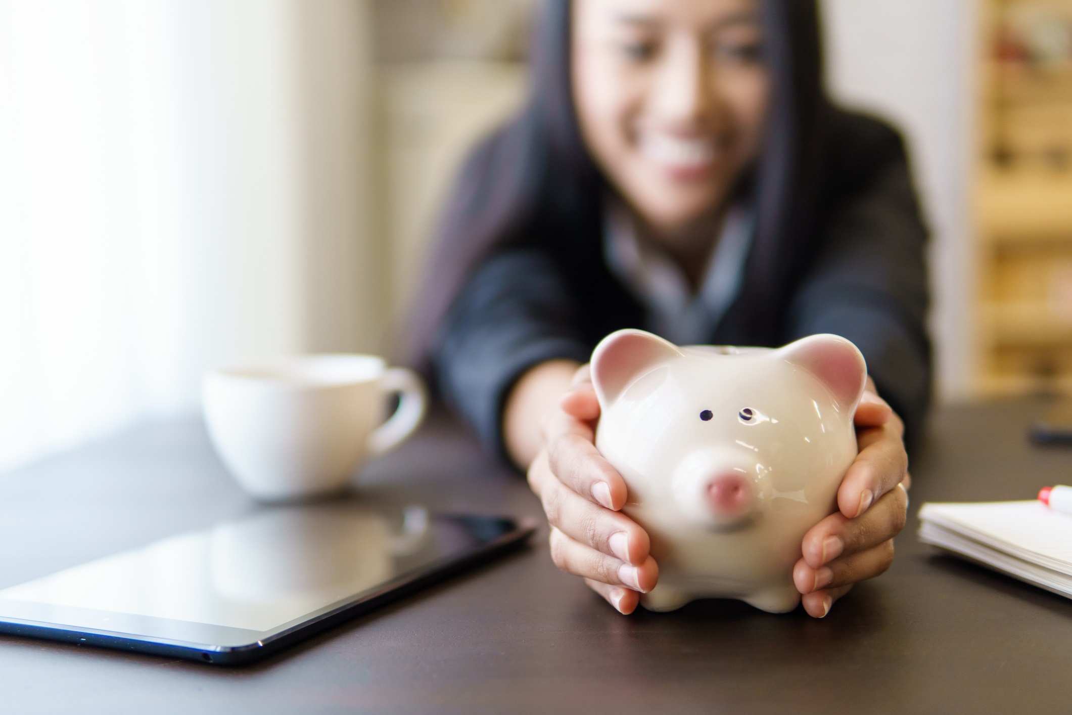 Female Teen Sitting at a Table Smiling and Holding a Coin Piggy Bank with a Tablet and Cup