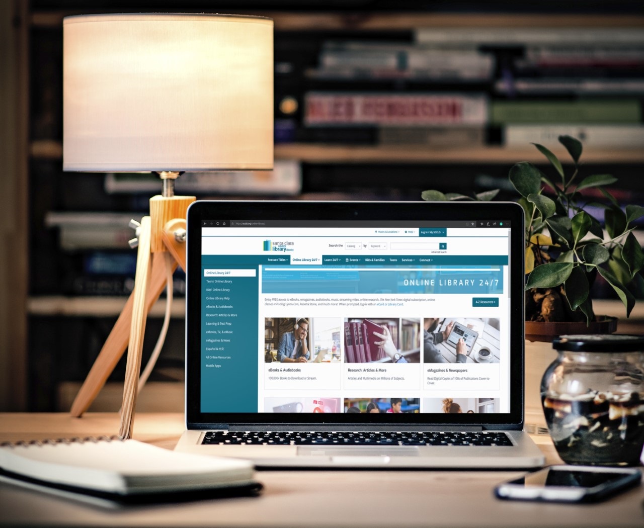 A laptop computer featuring SCCLD's Online Library landing page. The laptop is sitting on a desk next to a notebook, lamp and plant. 