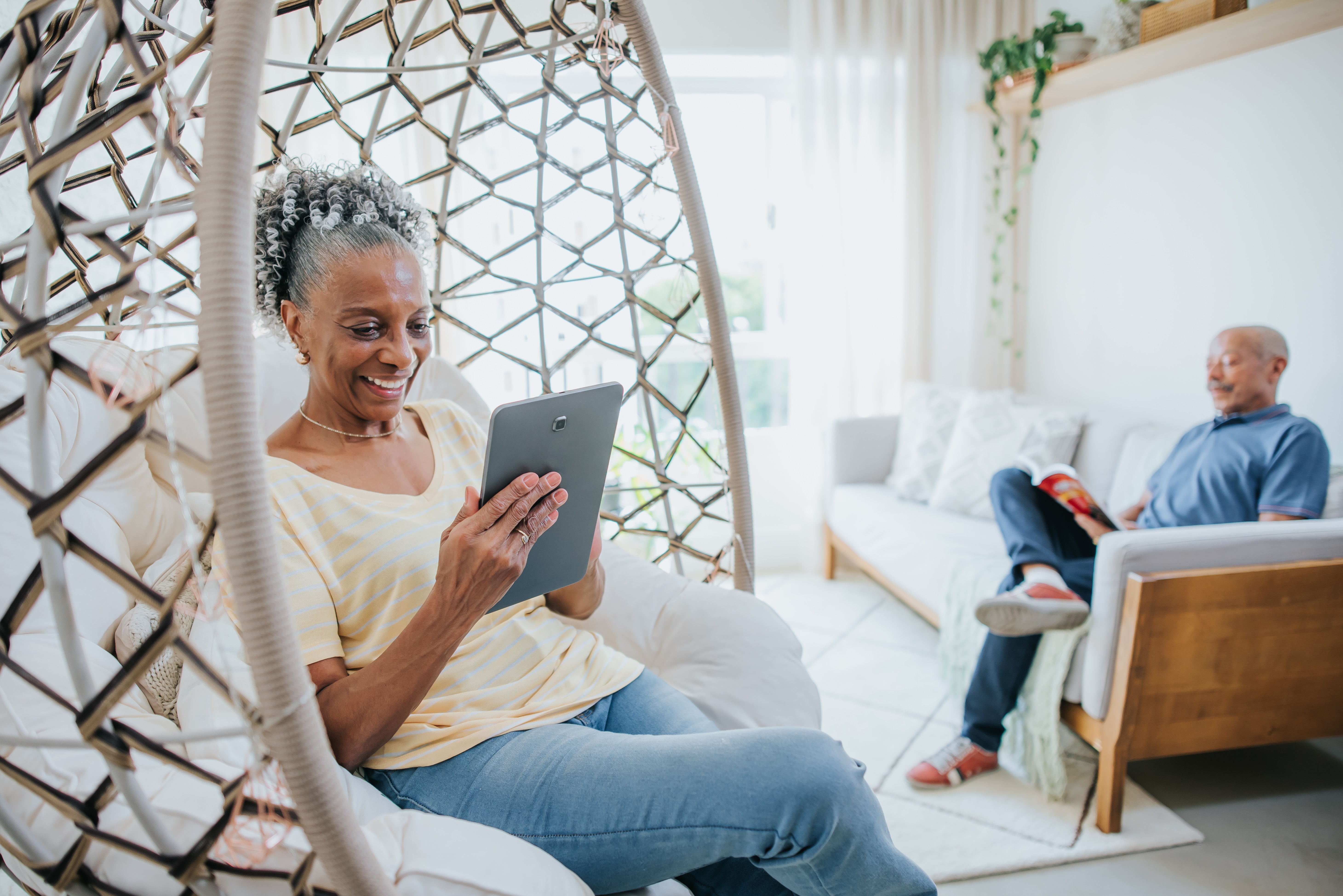 A mature adult sitting on a hanging chair using an e-reader. 