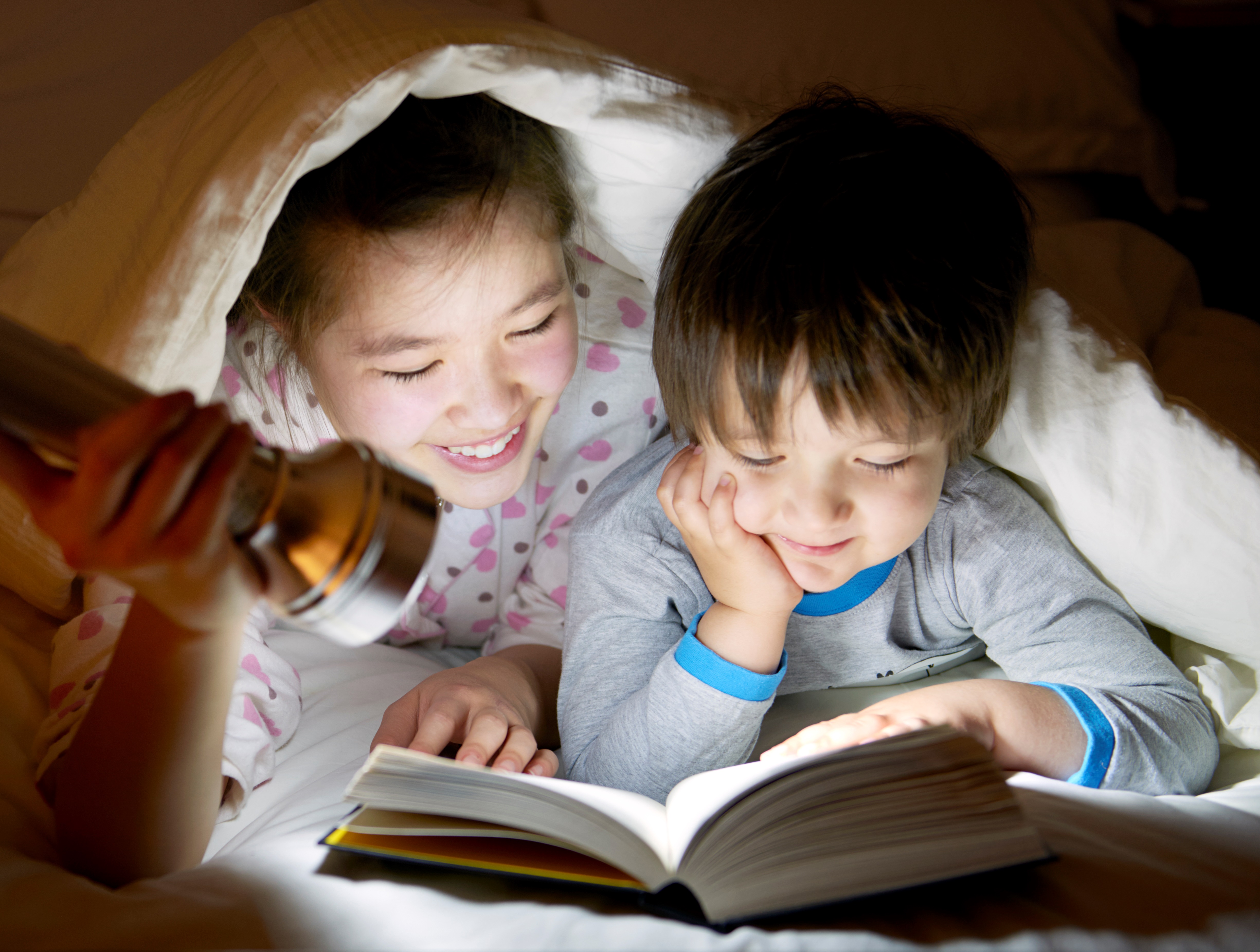 Siblings Reading a Book with a Flashlight under the Covers 