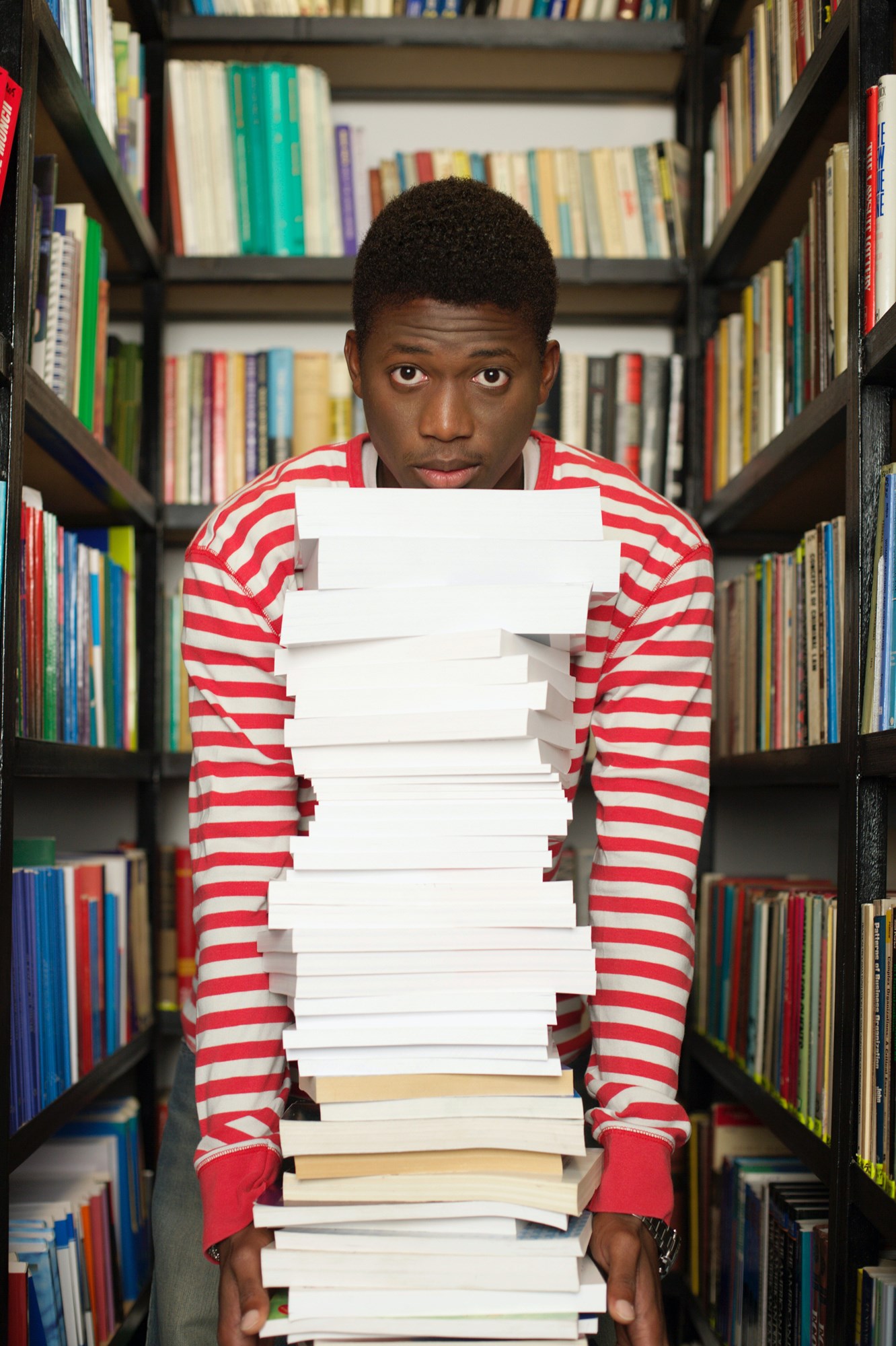 A person wearing a red and white shirt is standing among bookshelves, looking at the camera, and holding a large stack of books