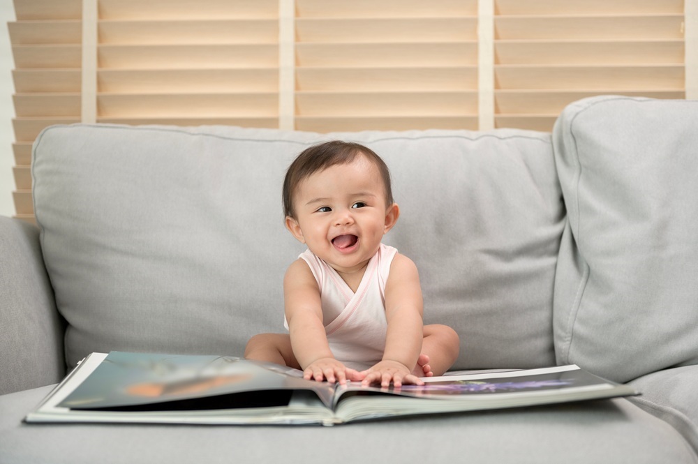 A cute baby girl sits on a gray sofa reading a large book.