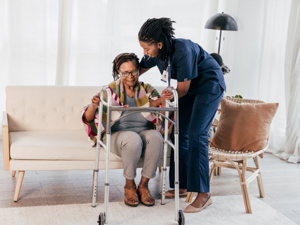 A nurse helping a lady with a walker stand up from sitting on a couch