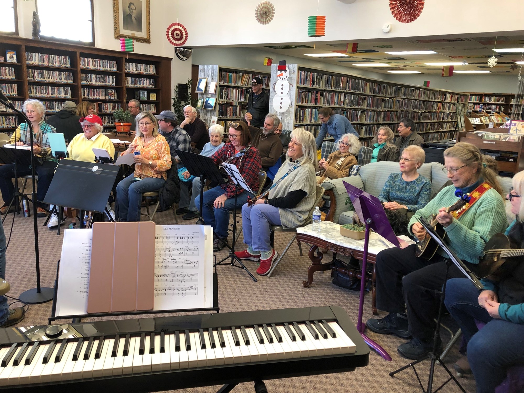 A large group of people sit in the main room of the Powers Memorial Library holding a variety of instruments, like guitars, ukuleles, drums. There are music stands in front of them and many look like they are singing. There is a keyboard in front of the group.