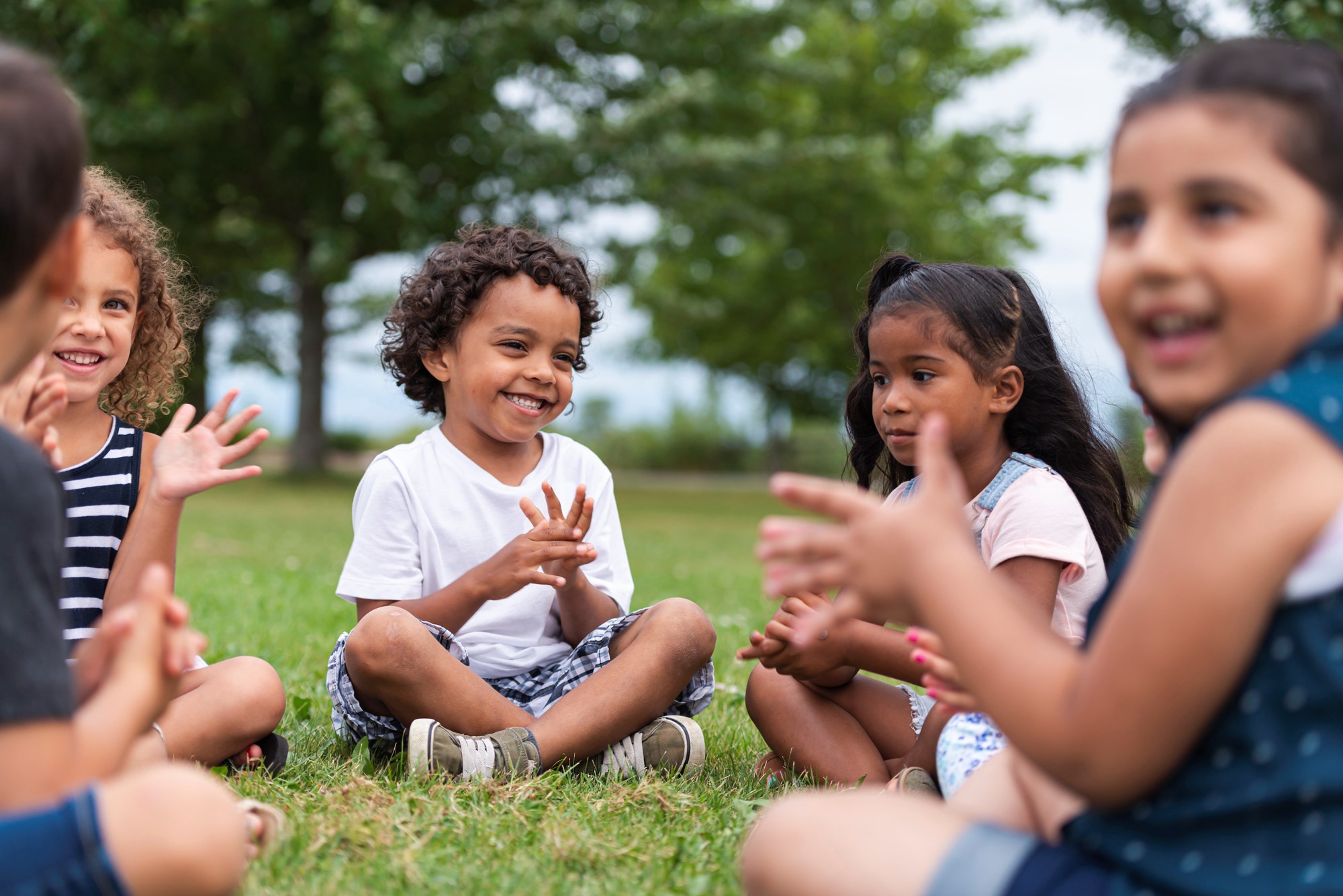Five children sitting on the grass outside smiling with each other.