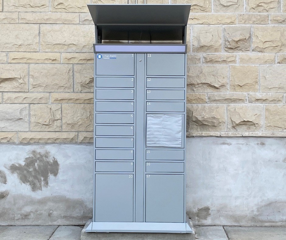 A tall gray smart locker standing outside a brick wall of the Waukesha Public Library. The locker is metal and looks like an outdoor postal box with drawers and an upper slot at the top.