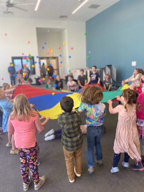 Children gather around a large colorful parachute with many balls flying up in the air 