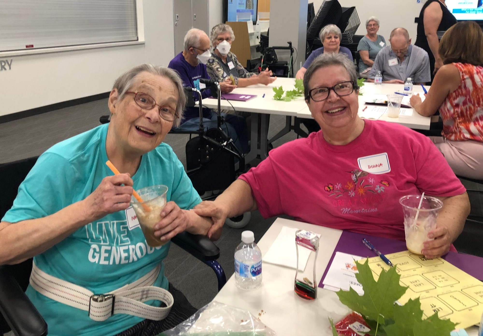 Two elderly people sit at a table holding plastic drink cups. They're attendees at a maple syrup program at the Library Memory Project. Plastic bottles, pieces of paper, and green maple leaves are on the table in front of them. A group of more elderly people are sitting at a table behind them.