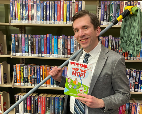 Pewaukee Public Librarian Peter Blenski holding a copy of 2026 Geisel Award Winner “Stop That Mop!” and a mop.