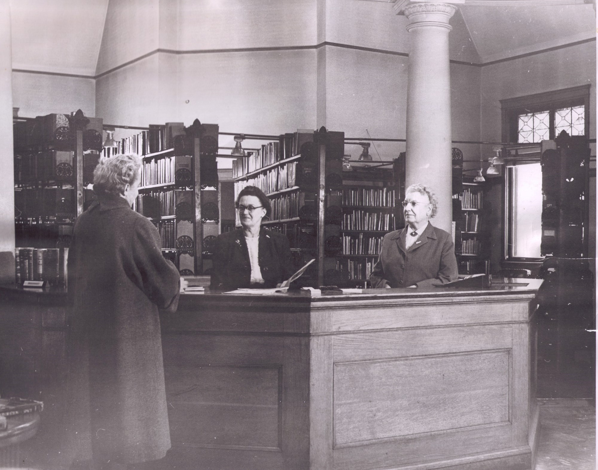 Two female librarians behind information desk and a female patron at desk talking to them; Picture from1952 at library building's 50th anniversary celebration
