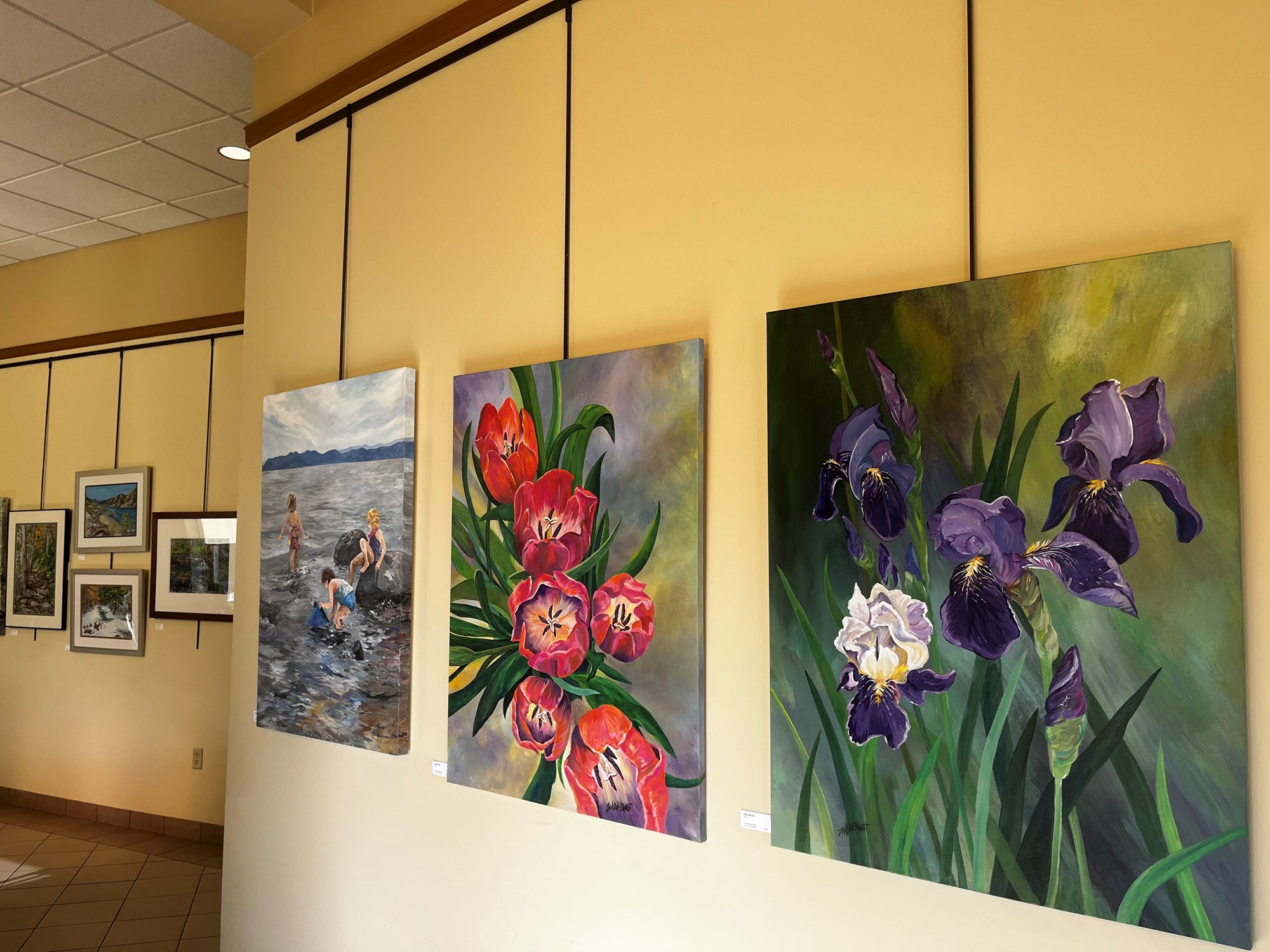 Two large canvas paintings of flowers and one of children swimming outside hanging in library's gallery