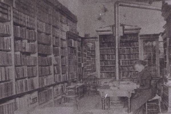 Black and white historical photo of a woman working at a desk in a library, surrounded by floor to ceiling shelves of books