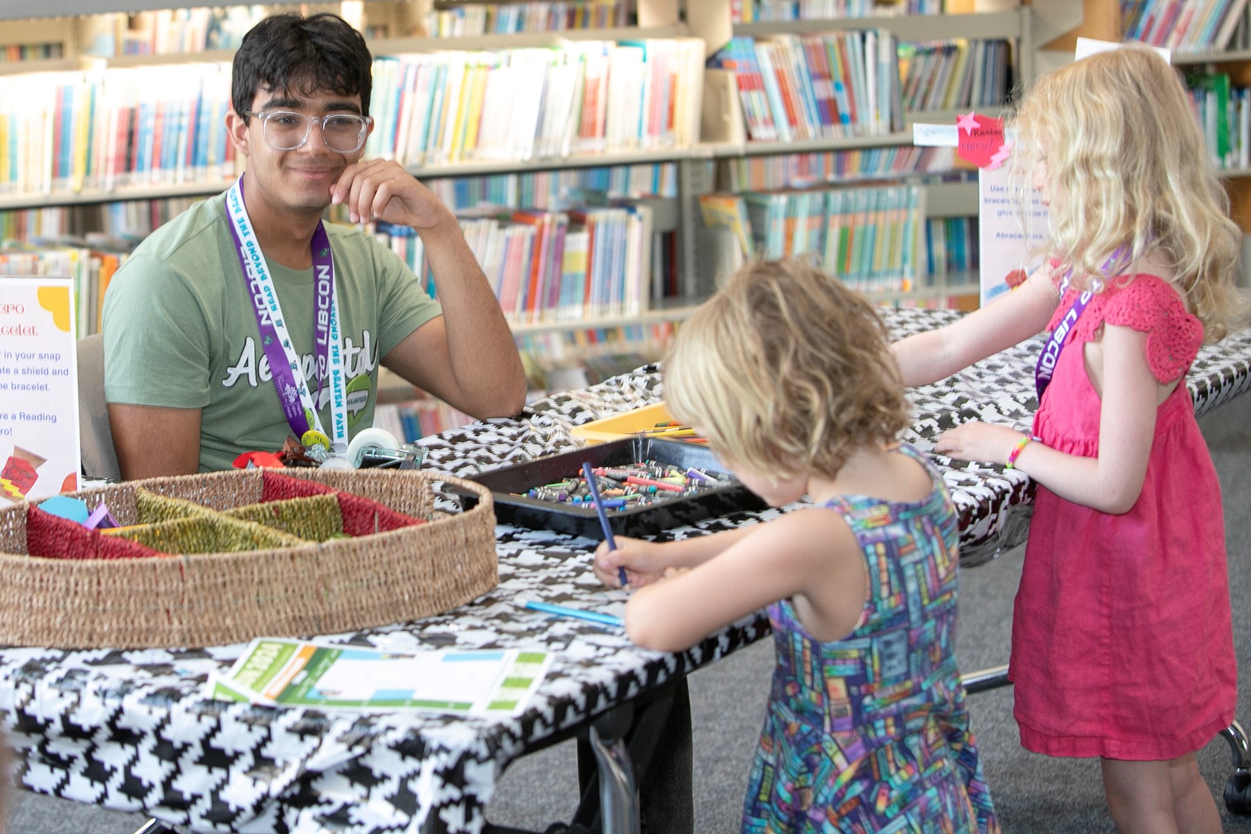 Teen volunteer watches kids color at summer reading table.