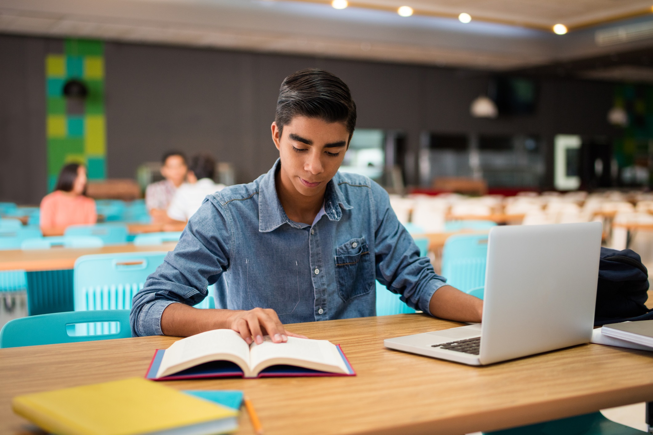 Student Sitting in Library with Laptop and Book 