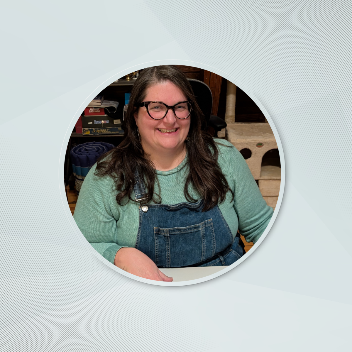 Author and Storyteller Katie Spina sits relaxed at a table in overalls and a mint green shirt. Her smile is welcoming.