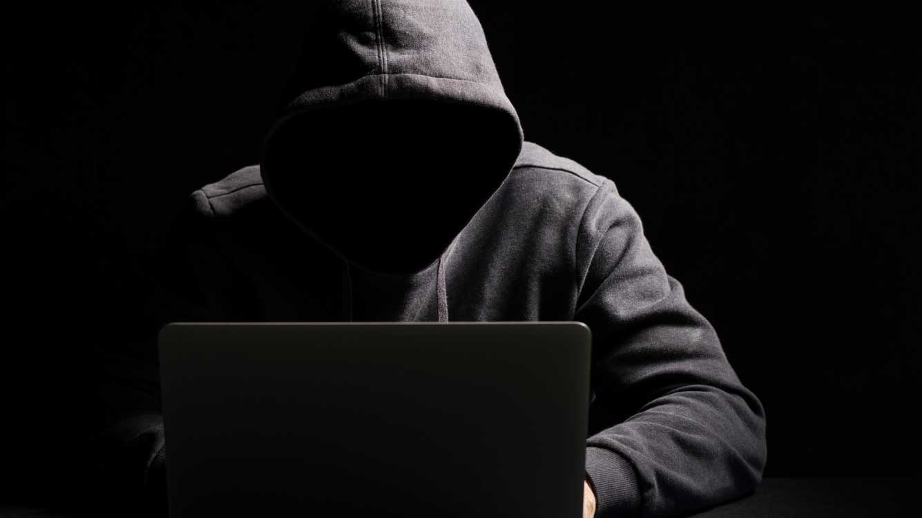 A hooded person with their face in shadow working on a laptop. A hooded person with their face in shadow working on a laptop.