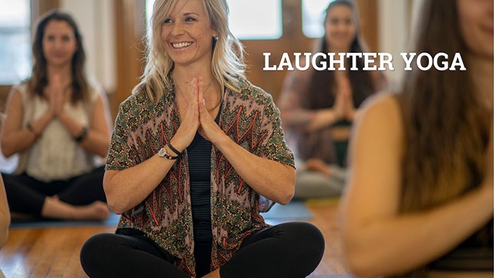 Group of people seated on yoga mats in a wooden-floored room, hands pressed together in a prayer position. Text on the image reads “LAUGHTER YOGA.”