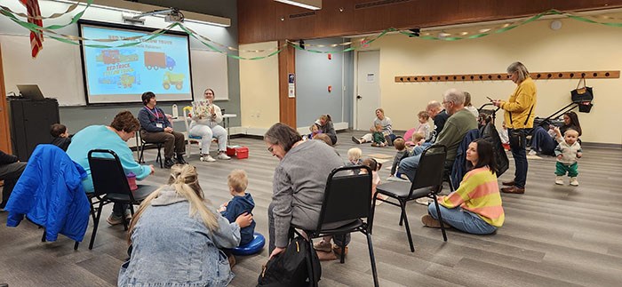 Adults and young children gather in a large meeting room for a storytime program. Participants sit on chairs and on the floor in a circle around two presenters at the front of the room. A projected slide is visible on a screen, and colorful streamers hang across the ceiling. Adults and young children gather in a large meeting room for a storytime program. Participants sit on chairs and on the floor in a circle around two presenters at the front of the room. A projected slide is visible on a screen, and colorful streamers hang across the ceiling.