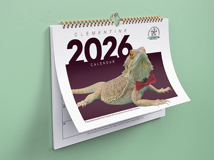 Wall calendar for the year 2026 titled 'Clementine 2026 Calendar.' The cover features a bearded dragon named Clementine, shown in a realistic photo wearing a bright red bow tie. Clementine is positioned diagonally across the page on a deep maroon background, with large white text displaying '2026' and smaller text reading 'Calendar' underneath. At the top, the name 'Clementine' appears in uppercase letters, and the Friends of the Oconomowoc Public Library logo is visible in the upper right corner. The calendar is spiral-bound and hangs against a light green wall. A portion of the next calendar page is visible beneath the cover.