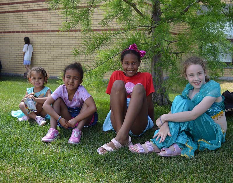 Four children sit on grass in the shade of a tree near a brick building; one holds a snack while another in a teal costume sits at right.