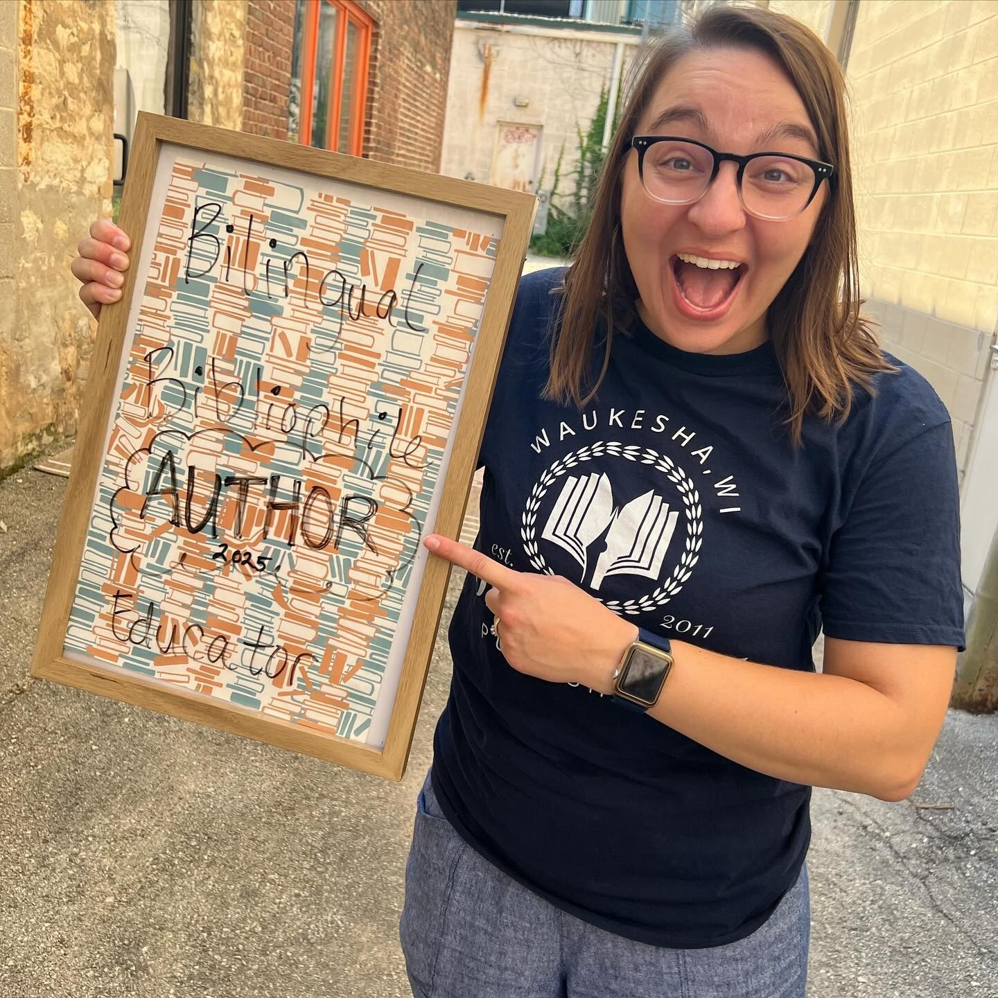 Author Amanda Stewart holding a sign that reads "Bilingual, Bibliophile, Author 2025, Educator."