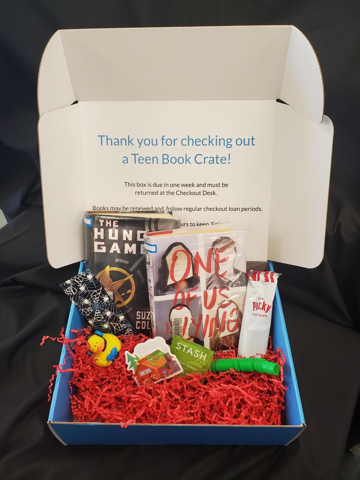 A teen book crate with The Hunger Games, One of Us Is Lying, snacks, small toys, and a thank‑you message displayed inside the box lid.