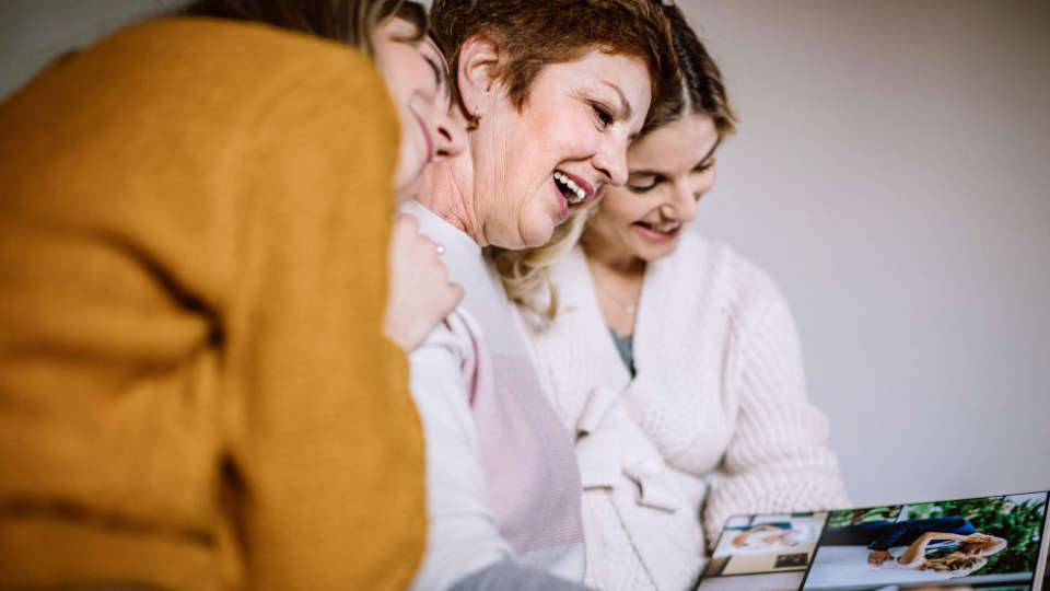 Three people sit closely together looking at a photo album or printed photographs. One person in the center turns a page while the others lean in to view the images. The photo is a close-up with a softly blurred background and warm indoor lighting.