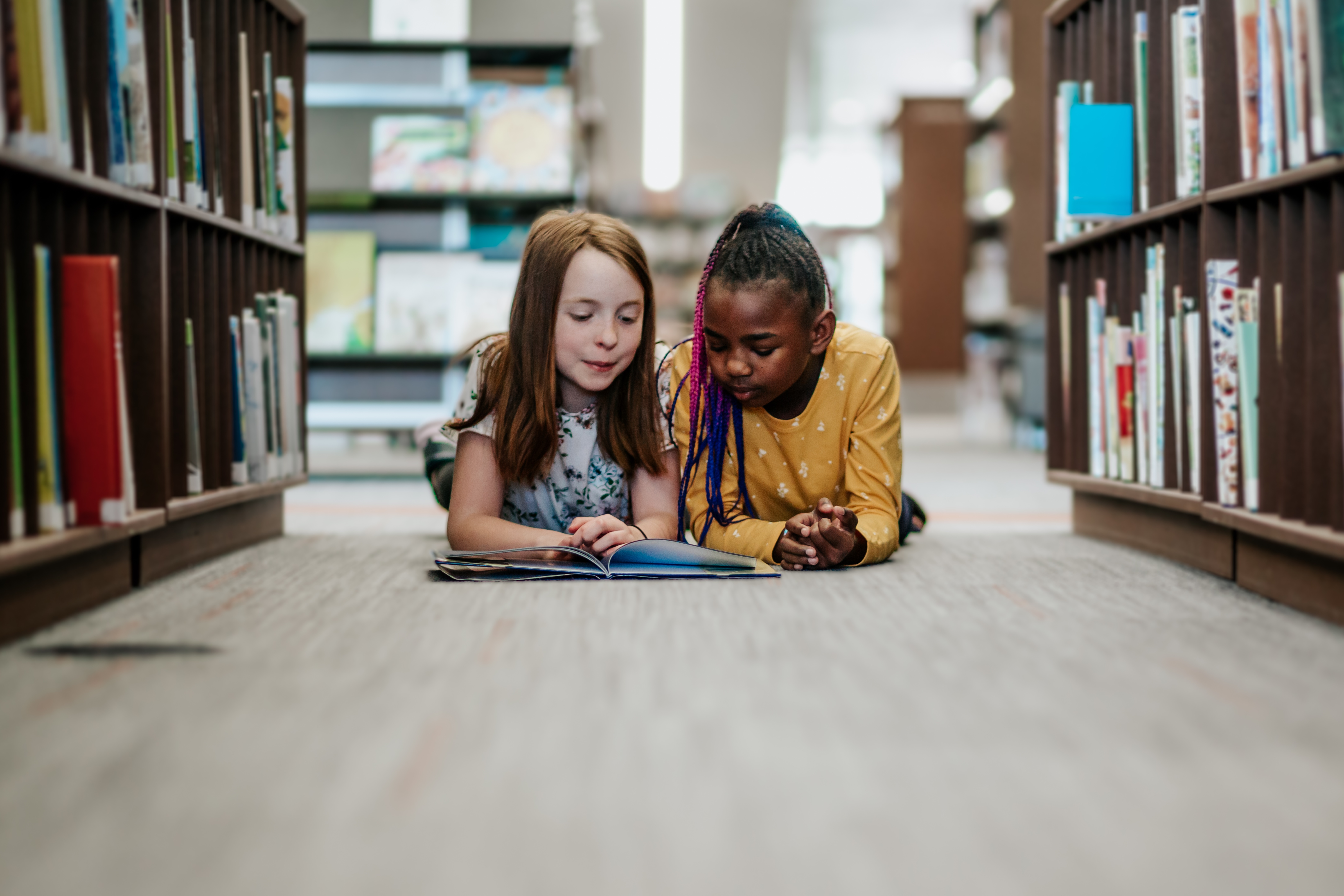 Two Young Girls Reading a Book on the floor of Library