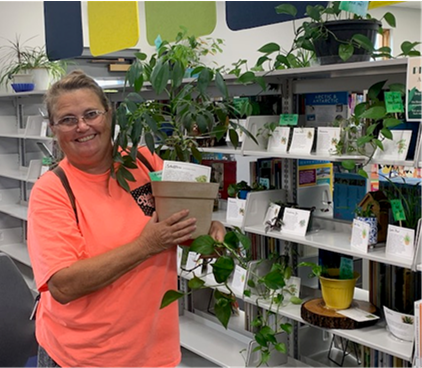 Smiling woman in an orange shirt holding a potted plant in front of shelves of plants