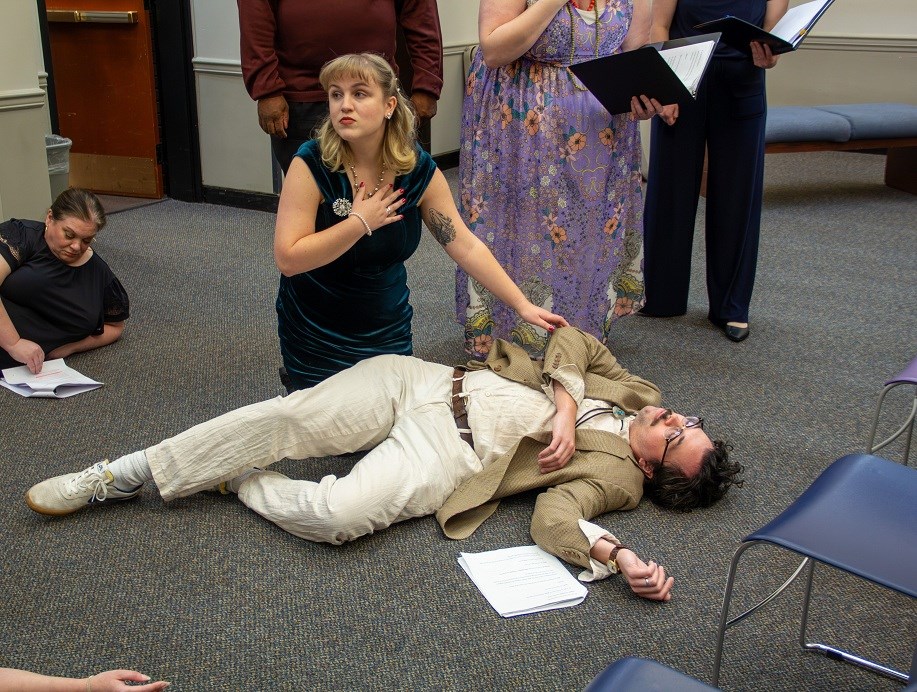 Woman clutching her heart while kneeling over a body of a man at a library murder mystery event
